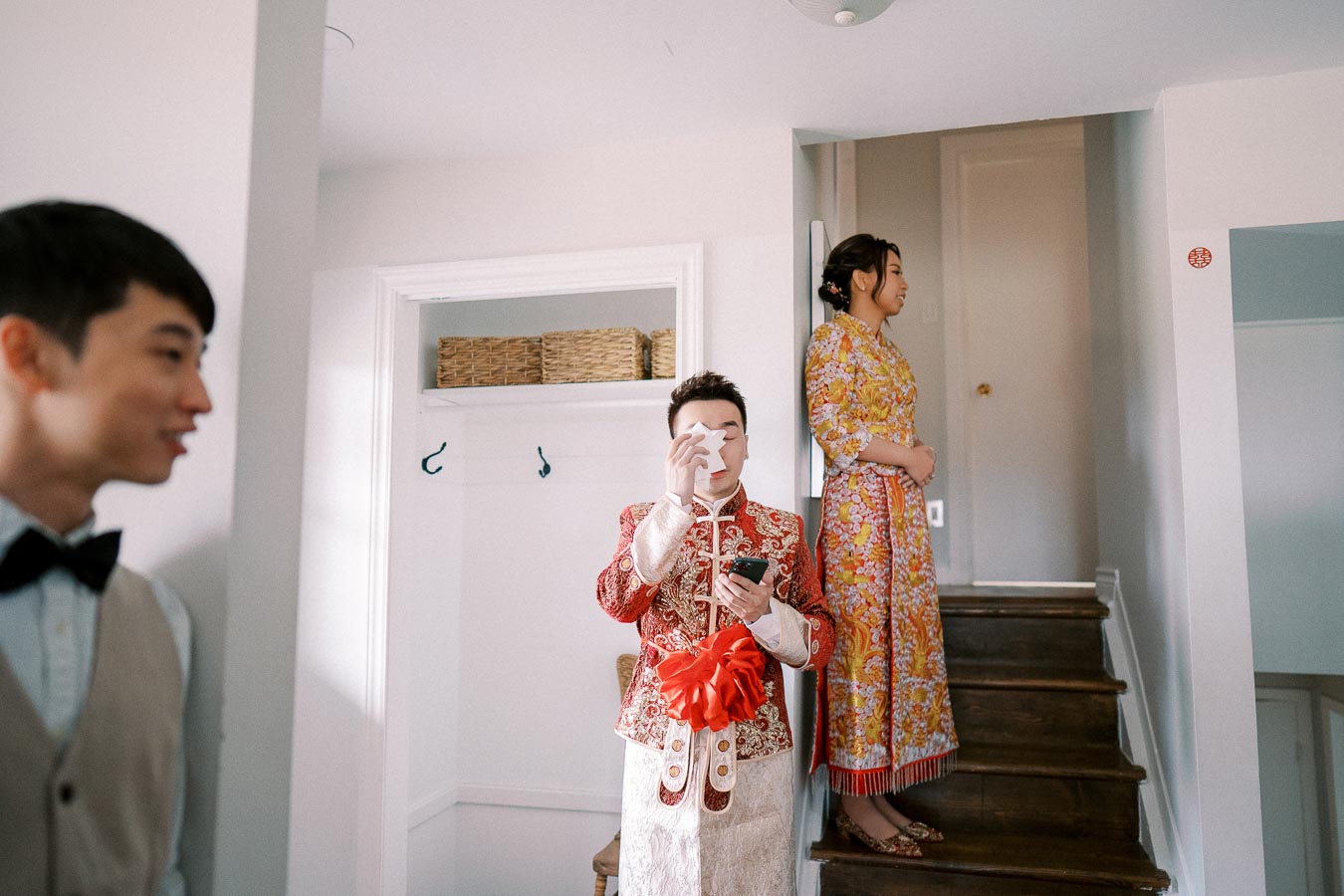 Groom in traditional embroidered Chinese attire prepares for wedding ceremony, while bride in colorful dress stands nearby on staircase, with another guest smiling in the foreground.