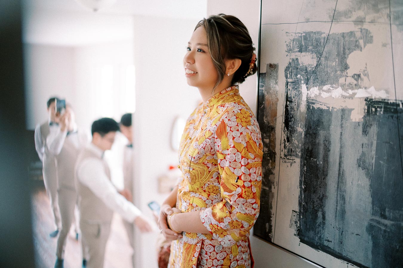 Woman in floral dress smiling indoors, with abstract artwork behind her. Other people in background capturing the moment with a smartphone.