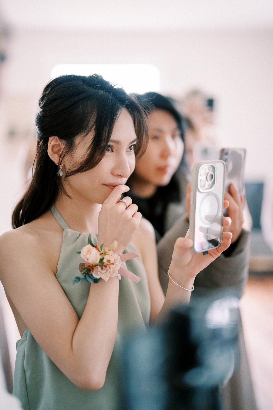 Woman in a sleeveless dress with a floral corsage, capturing a moment on her smartphone, surrounded by soft natural lighting.