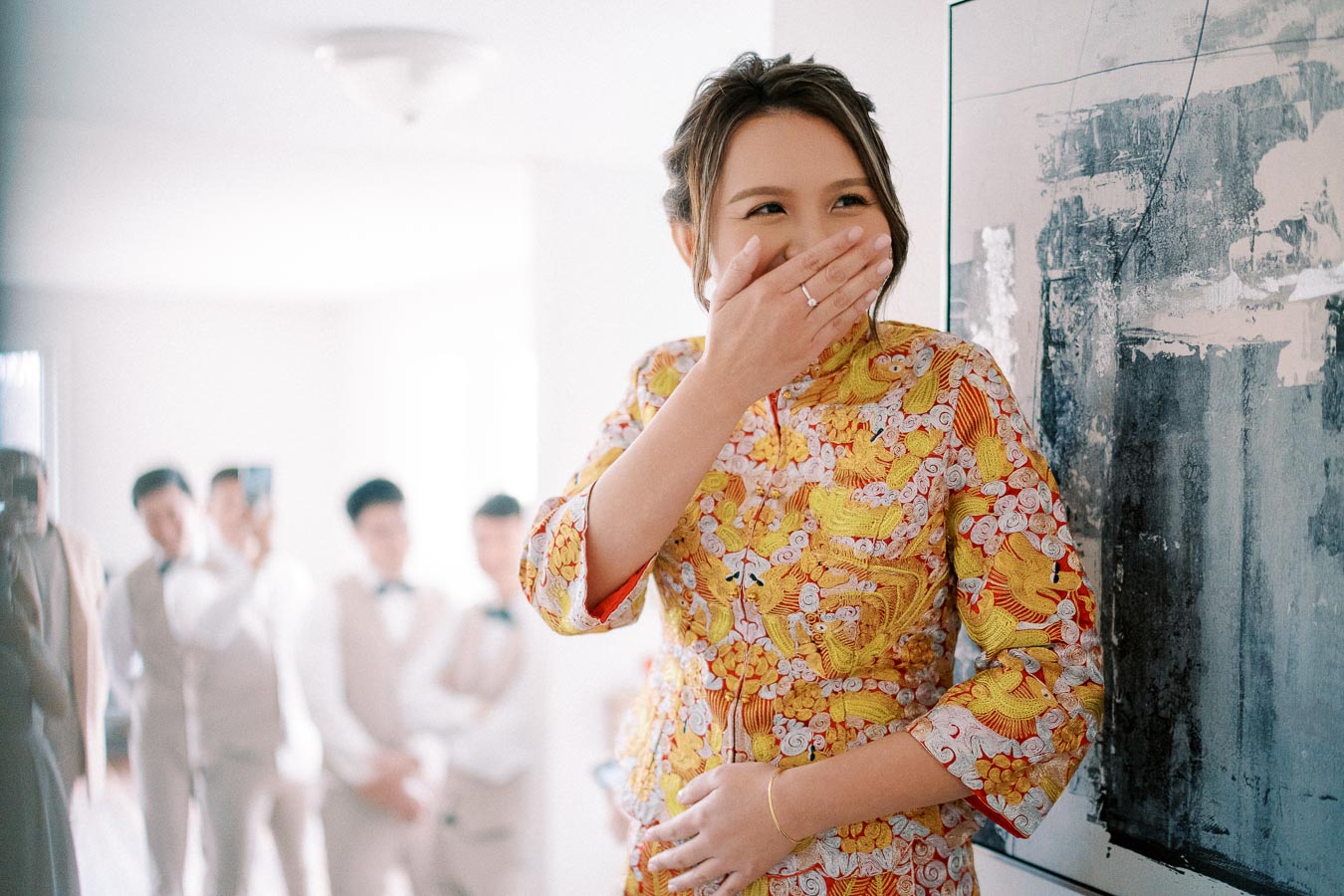 A woman in a colorful traditional outfit smiles, covering her mouth with her hand, standing in a bright room with blurred people in the background, capturing a candid and joyful moment.