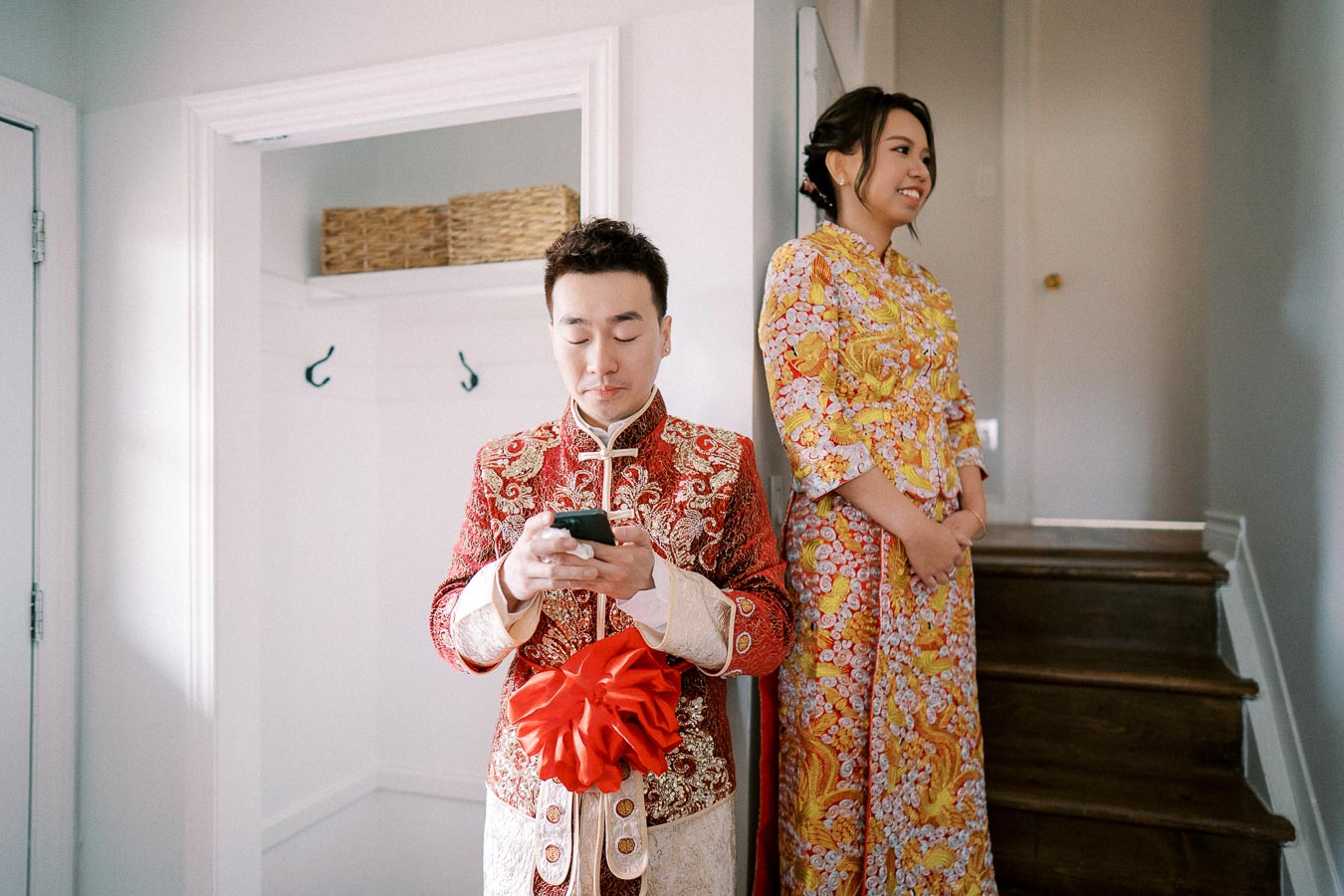 A couple dressed in traditional Asian attire during a wedding ceremony; the man types on a smartphone while wearing an ornate red and gold outfit, and the woman smiles in a vibrant yellow and orange dress.