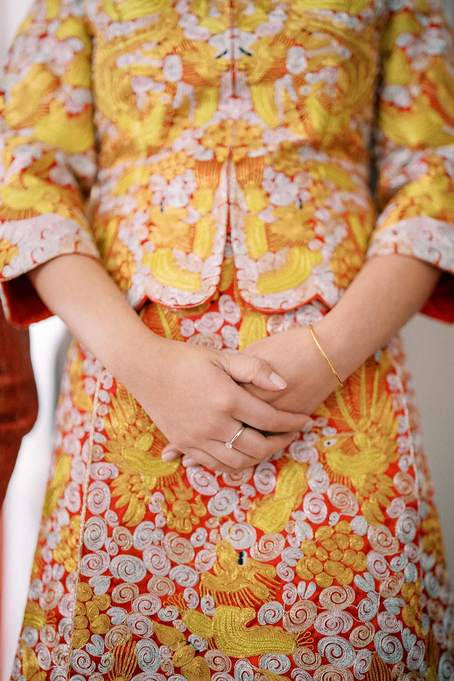 Close-up of a person wearing a vibrant traditional embroidered garment in red, yellow, and gold, showcasing intricate patterns and designs. Hands are clasped at the front, with a delicate ring on one finger and a thin gold bracelet on the wrist. Ideal alt text for cultural attire or fashion content.