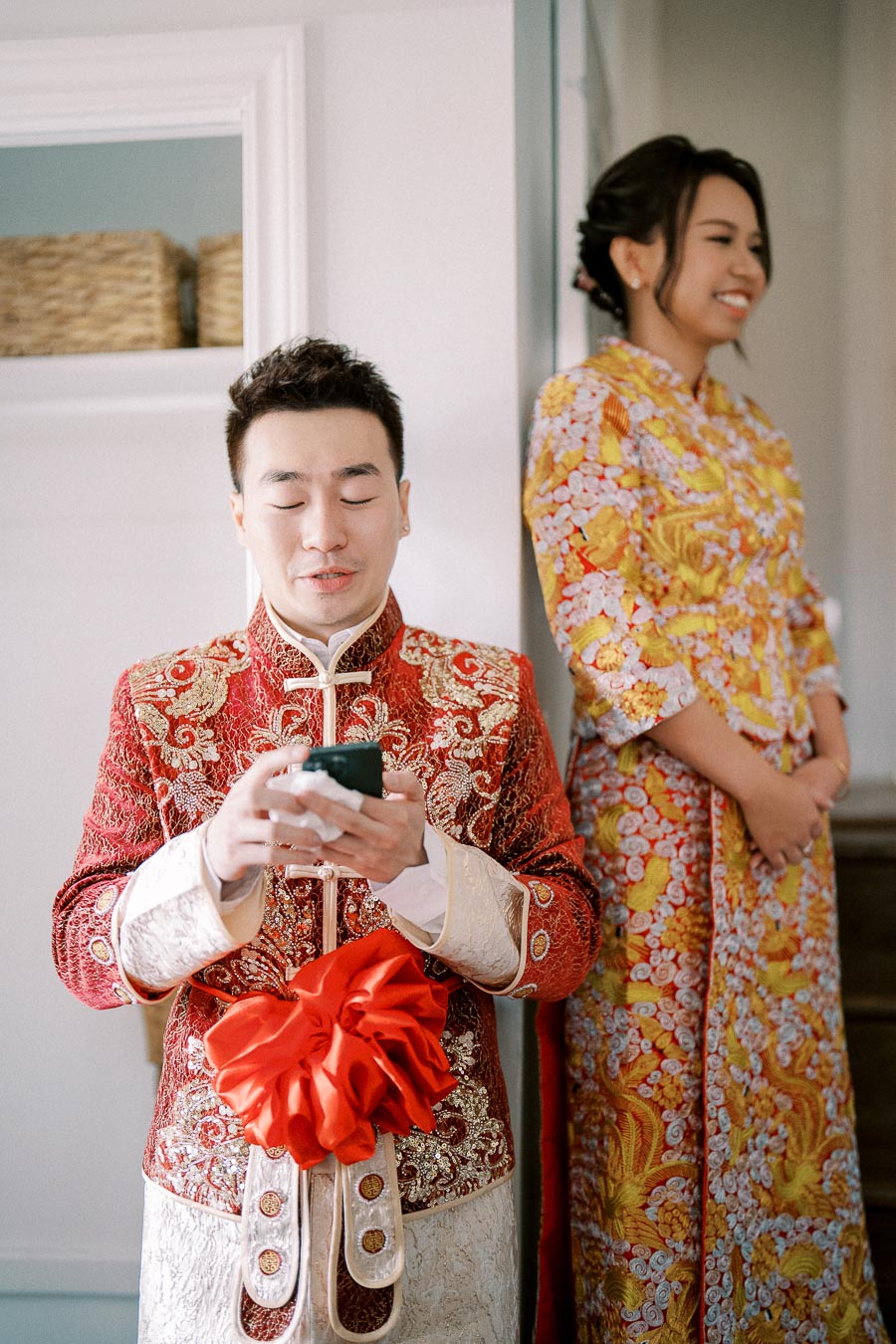 A man in traditional Asian wedding attire holds a phone, while a woman in a colorful embroidered dress stands nearby, both smiling, possibly preparing for a wedding ceremony.
