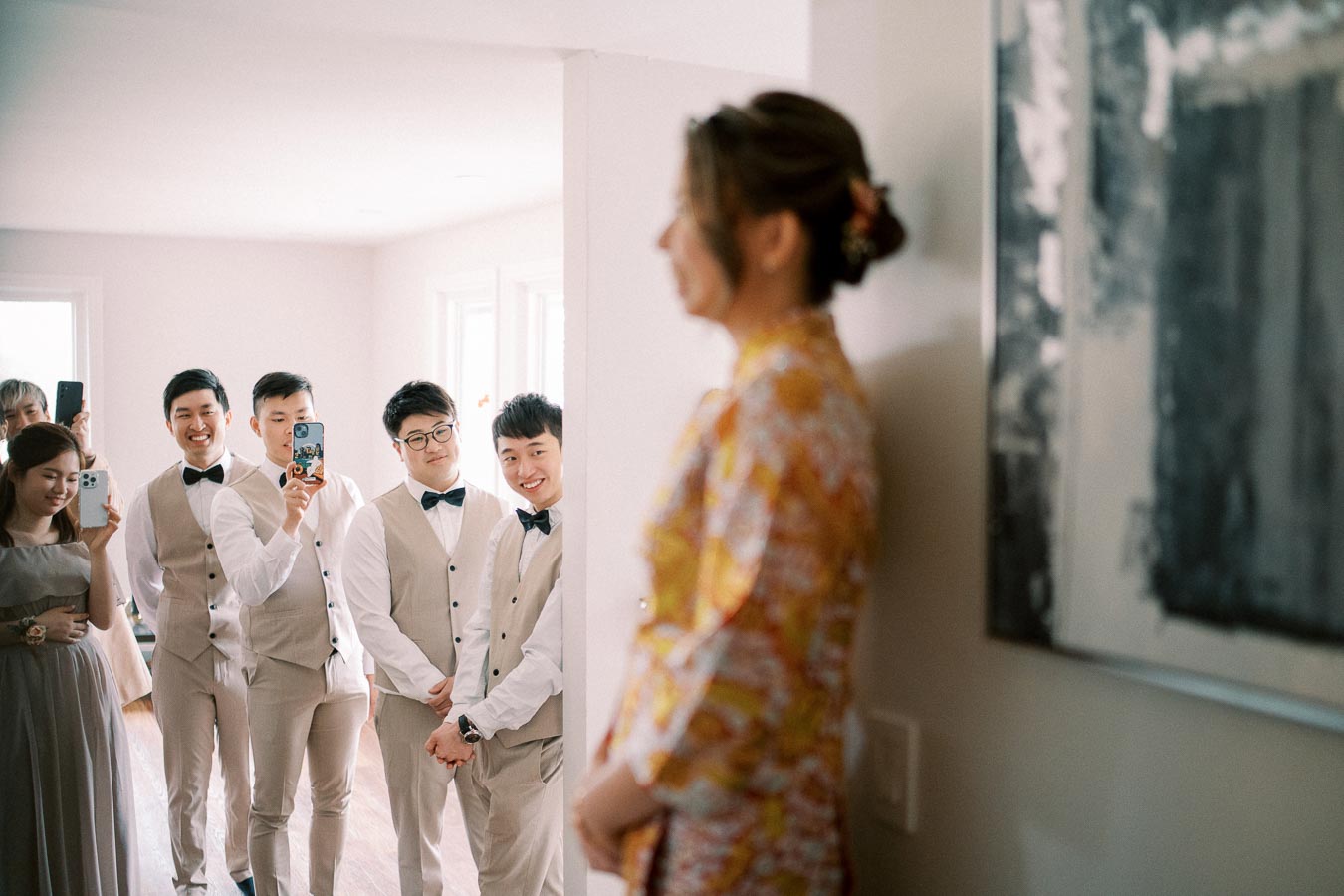 A group of groomsmen in beige suits with bow ties, smiling and capturing a moment with their phones, as they stand opposite a woman in a colorful dress, possibly during a pre-wedding gathering in a bright, sunlit room.