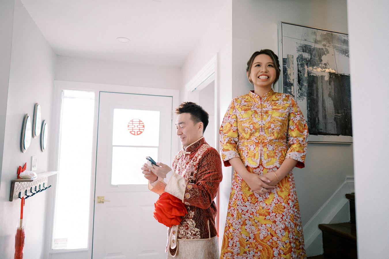 A couple wearing traditional Chinese wedding attire, with the bride in a yellow floral dress and the groom in a red embroidered outfit, standing indoors near a door adorned with a Chinese character.