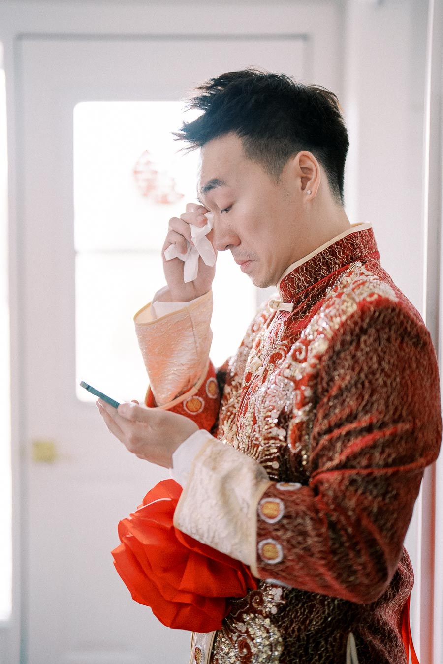 A groom in traditional Chinese wedding attire wipes a tear while holding a smartphone, capturing an emotional moment before the ceremony.