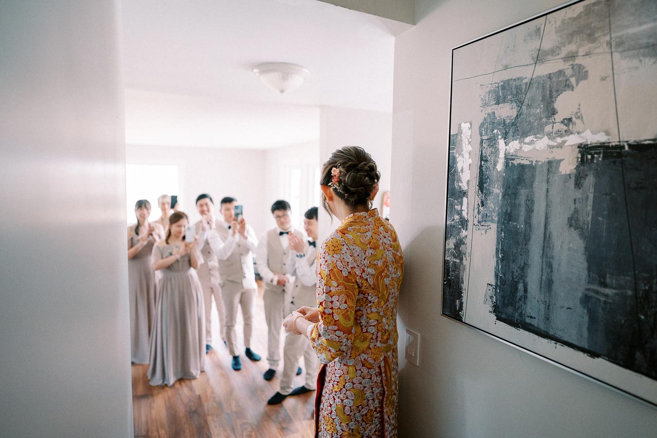 Bride in traditional Asian robe greets group of elegantly dressed guests inside a modern home, capturing a joyful wedding moment.