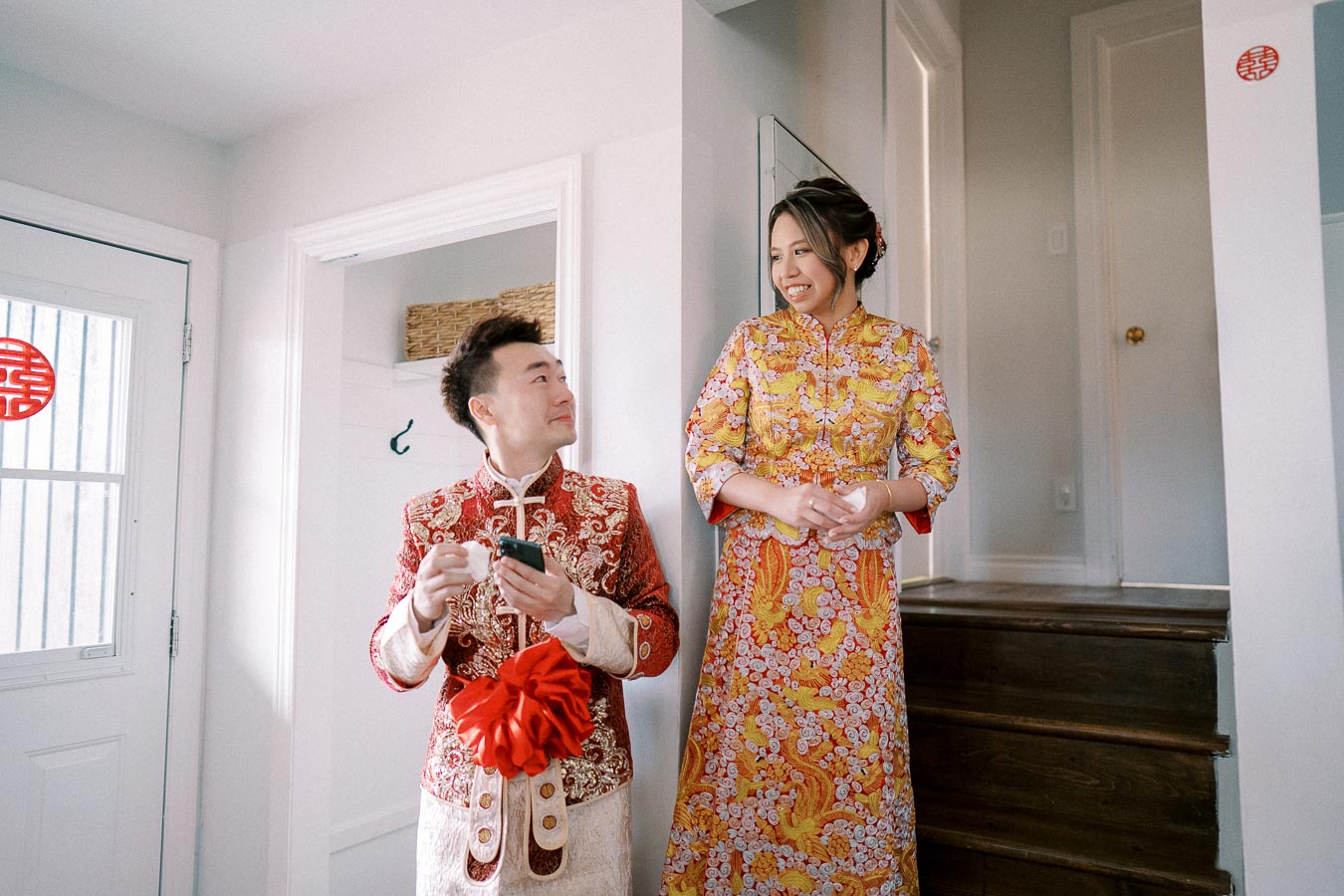 Couple wearing traditional Chinese wedding outfits, smiling at each other in a brightly lit home environment.