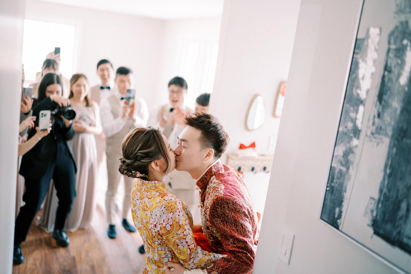 A couple dressed in traditional wedding attire shares a kiss while guests capture the moment on their cameras in a bright, joyful setting.
