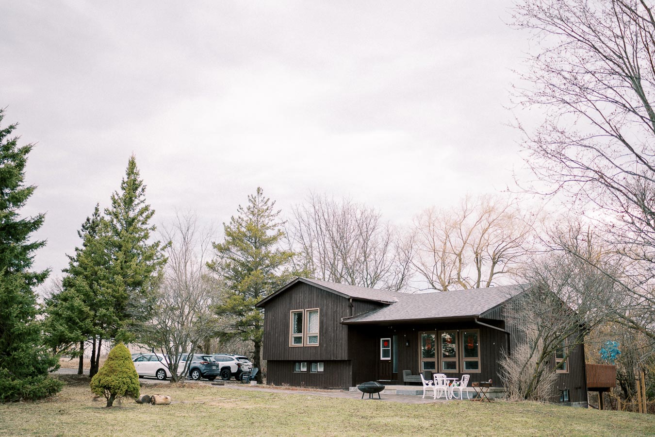 A cozy, modern brown house surrounded by lush green trees and a spacious yard, featuring an outdoor patio with white furniture, set under a cloudy sky.