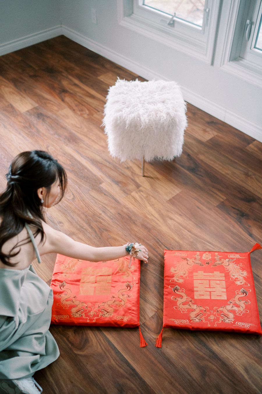 A woman arranging two red embroidered cushions with traditional Chinese symbols on a wooden floor, next to a fluffy white stool by a window.