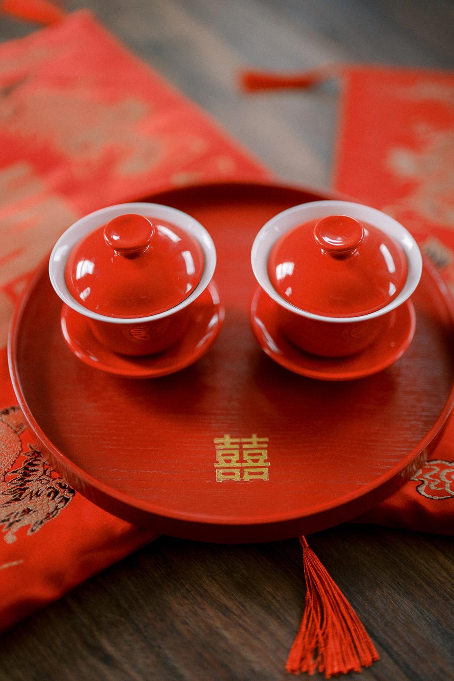 Red traditional Chinese tea set on a wooden table, featuring two lidded cups on a round tray with a golden double happiness symbol, used in wedding ceremonies.