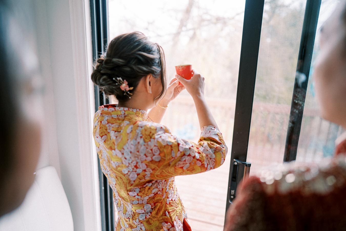 Person holding a red tea cup in traditional floral outfit near a window, showcasing cultural celebration