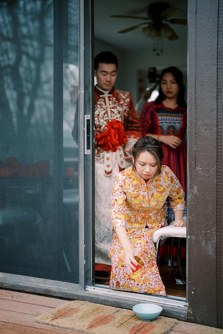 Traditional Asian wedding ceremony with a woman in an ornate dress pouring tea, accompanied by two people in traditional attire, outside a sliding glass door.
