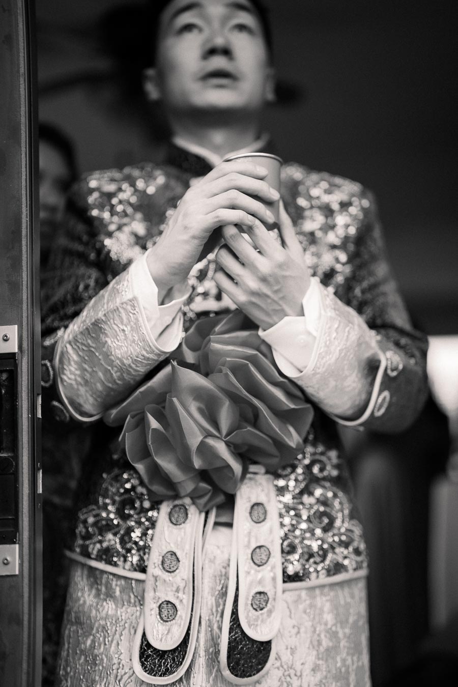 A person wearing an elaborate, embroidered garment holds a cup in both hands, standing indoors. The image is in black and white, highlighting the intricate details of the outfit and the serene expression on the person's face.
