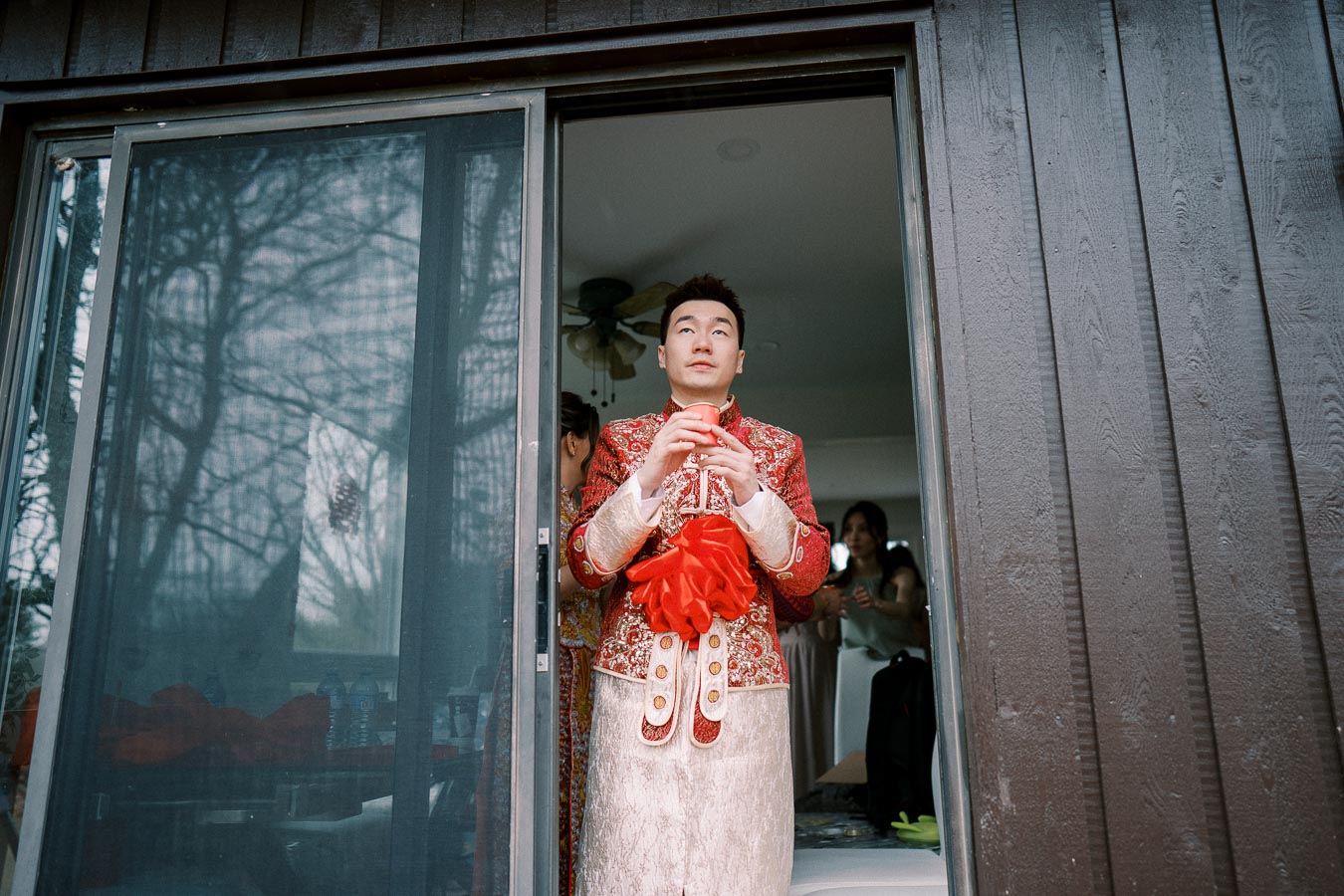 A person in traditional red and white attire holding a red ribbon stands in a doorway, with a partially open sliding glass door reflecting trees on the left.