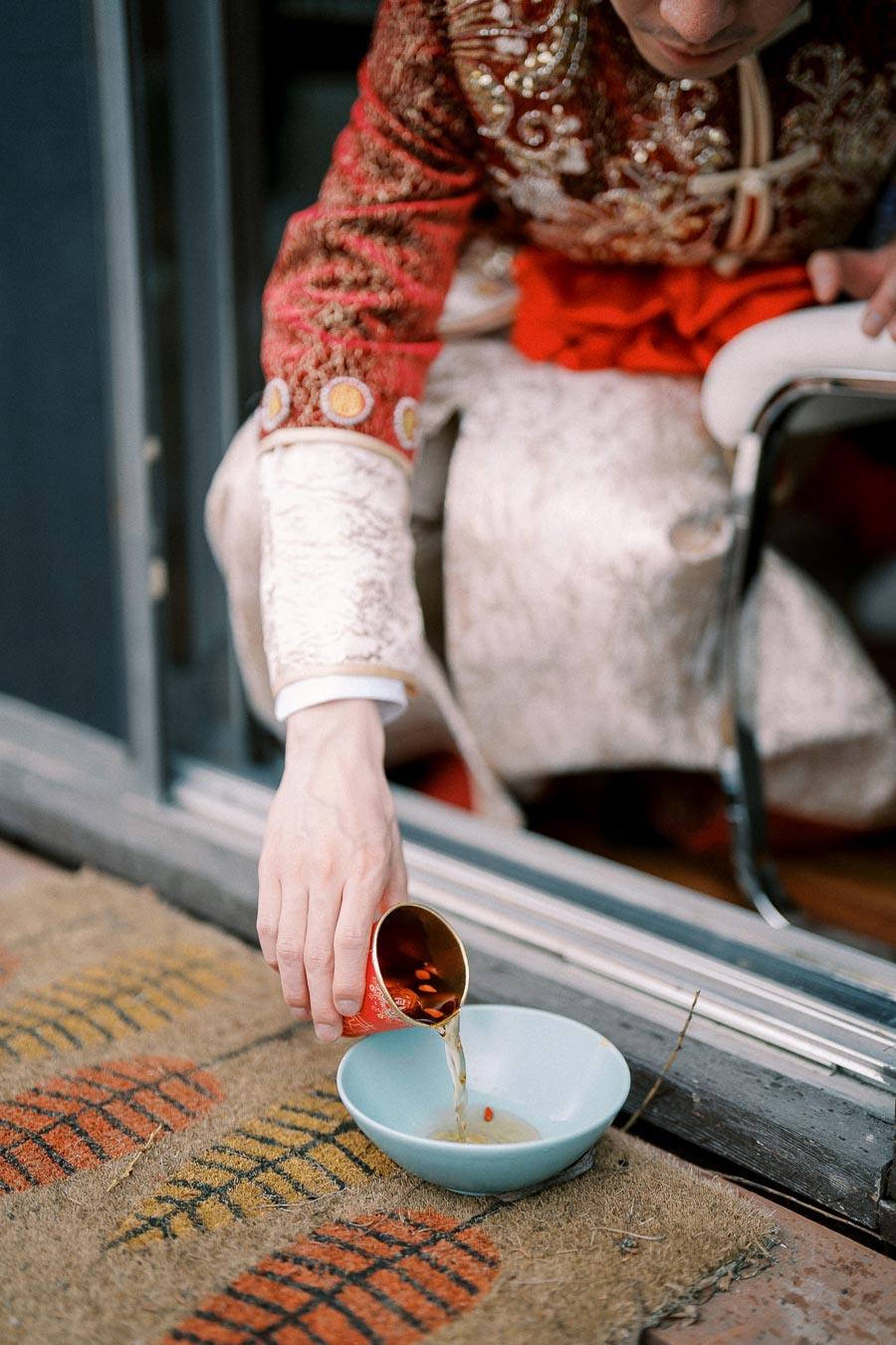 A person in ornate traditional attire pouring liquid from a decorated cup into a blue bowl placed on a patterned doormat.
