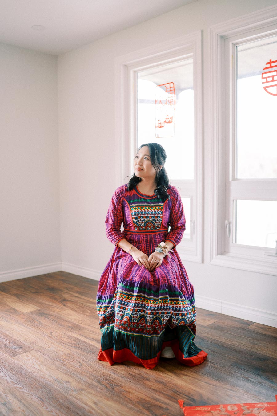 Woman in colorful traditional dress sits on a wooden floor, looking thoughtfully towards the window decorated with red symbols, in a bright and minimalistic room.