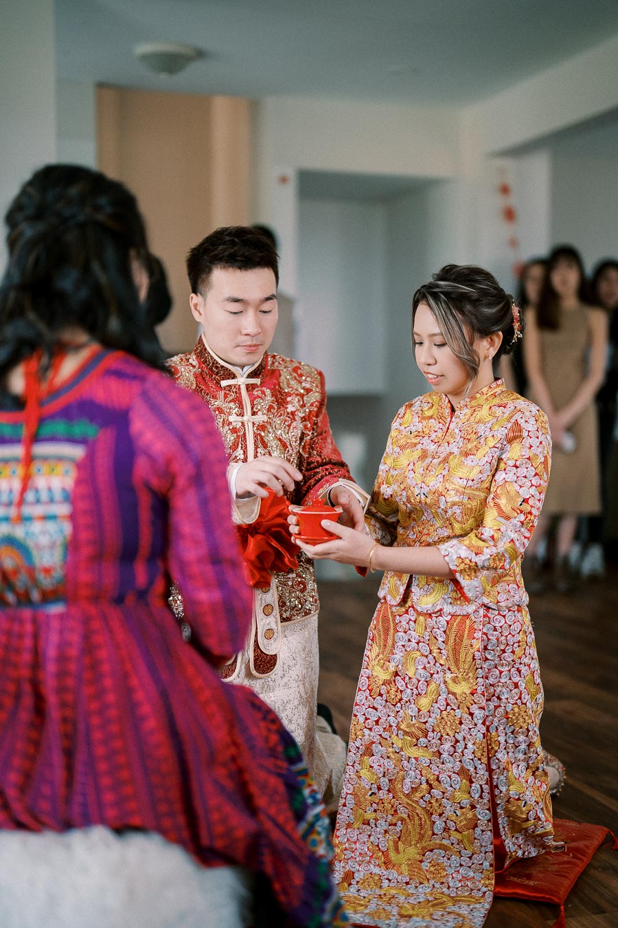 Couple in traditional attire participating in a Chinese tea ceremony during a wedding, with vibrant red and gold embroidery details.