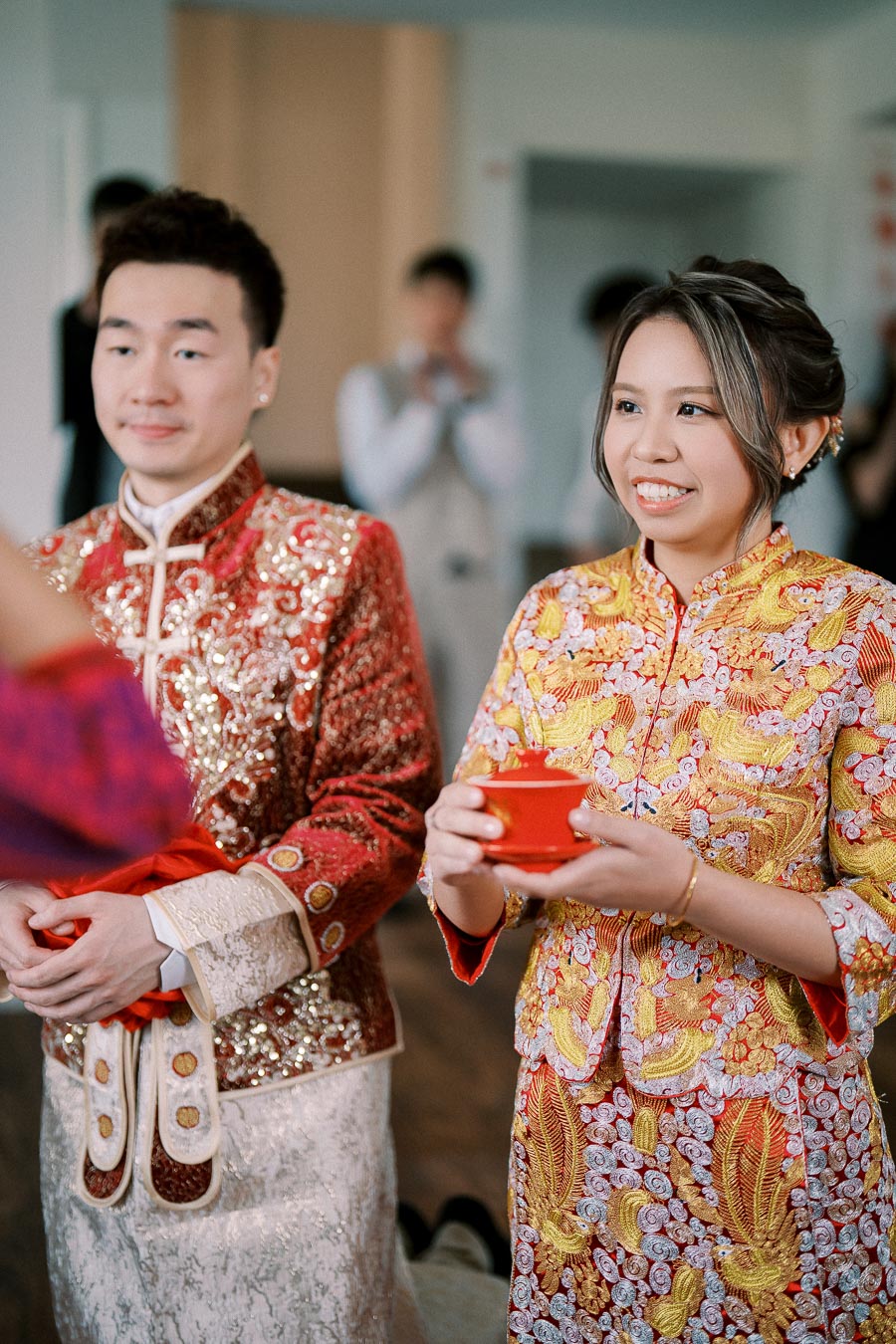 A couple dressed in traditional Chinese wedding attire during a tea ceremony, holding a red tea cup, symbolizing cultural heritage and celebration.