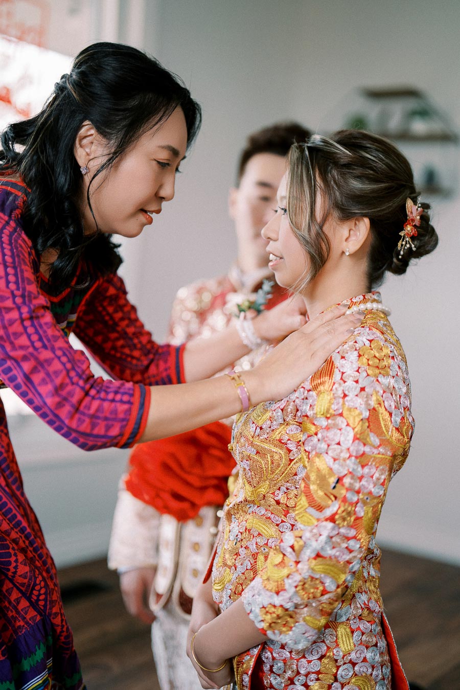 A woman dressed in colorful traditional attire adjusts the necklace of a bride in a vibrant, embroidered gown during a wedding ceremony.