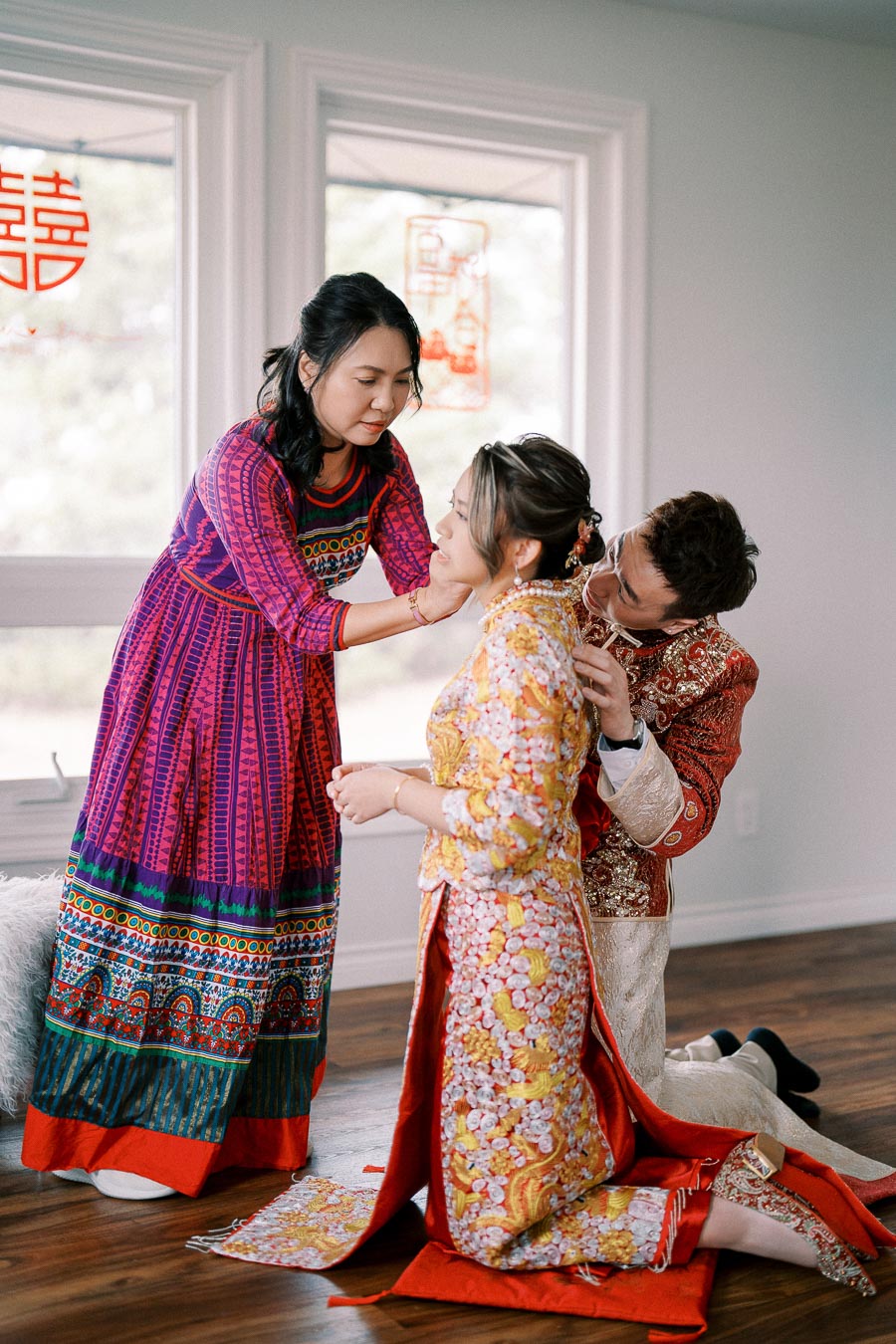 A bride in a colorful traditional Asian dress is assisted by two individuals, with red wedding decorations in the background, symbolizing a vibrant cultural ceremony.