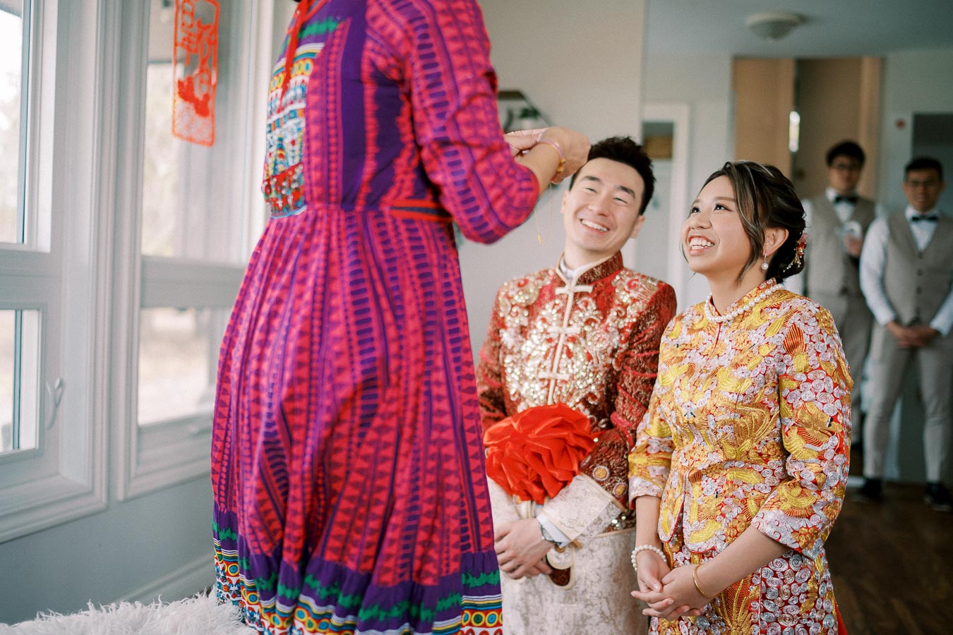 A couple dressed in traditional Asian wedding attire participating in a cultural ceremony, smiling as they look at a person wearing a colorful dress.