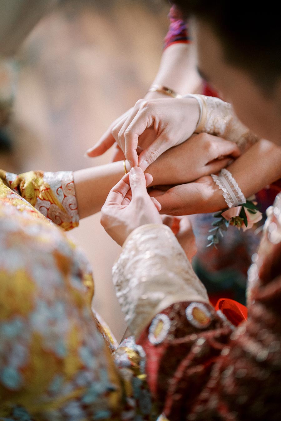 Hands delicately fastening a bracelet during a traditional ceremony, highlighting intricate, colorful attire and a floral bracelet in a close-up view.