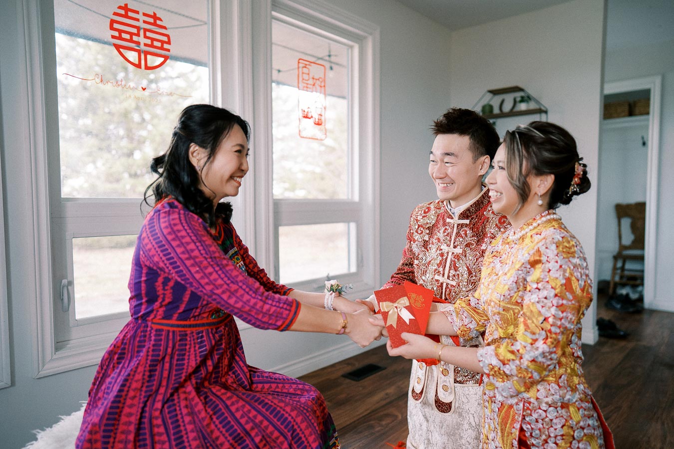 A happy couple in traditional Chinese wedding attire exchanging red envelopes with a seated woman in a vibrant dress during a wedding ceremony, with festive decorations in the background.