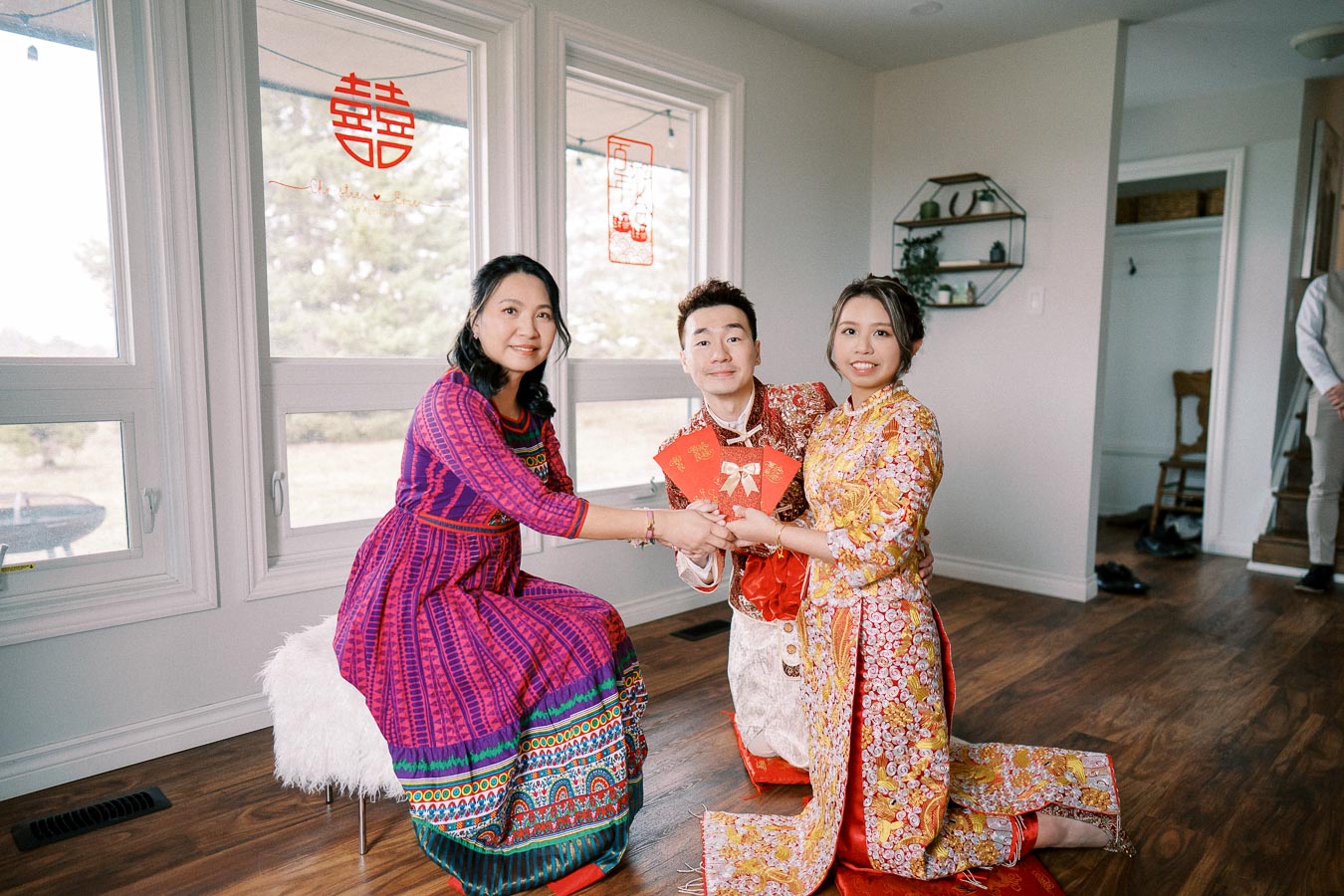 A couple in traditional Vietnamese wedding attire receives red envelopes while kneeling inside a decorated room, with festive symbols visible on the window behind them.