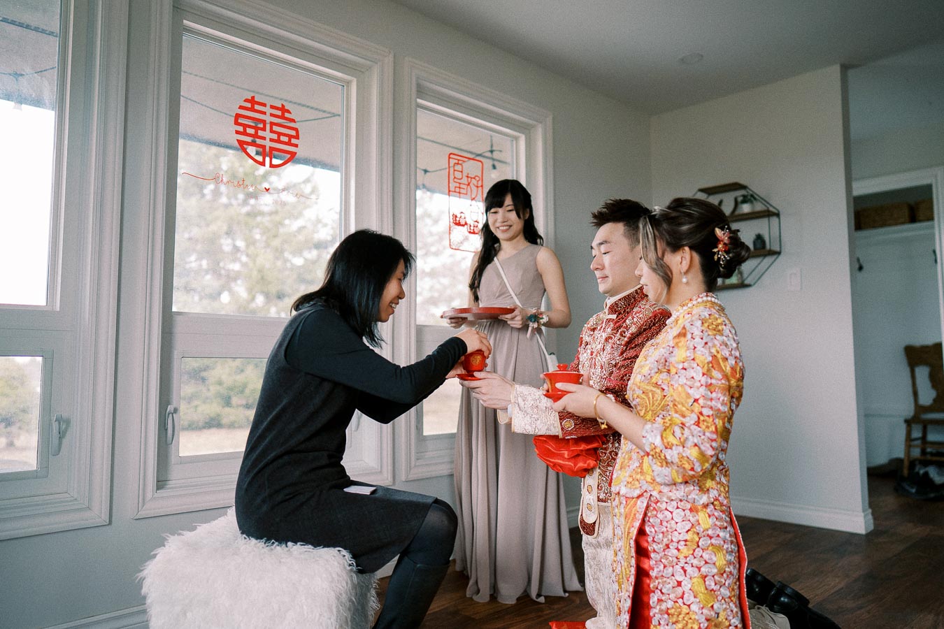 A group of people participates in a traditional tea ceremony inside a room with large windows. The individuals are dressed in formal attire, with vibrant red and gold traditional clothing, and the windows display red Chinese symbols.