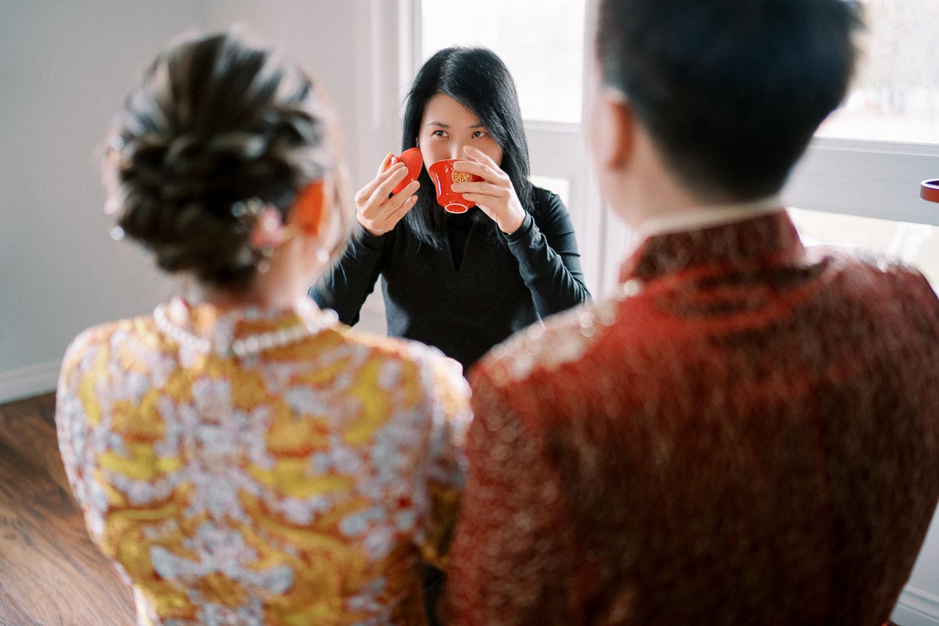 Asian woman in traditional clothing performing a tea ceremony, facing a couple dressed in ornate attire, symbolizing cultural traditions and celebrations.