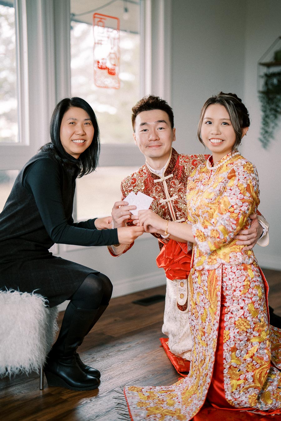 Group of three people participating in a traditional ceremony; two individuals in vibrant red and gold attire, representing cultural clothing, exchange envelopes with a woman dressed in black, sitting indoors in front of a bright window.