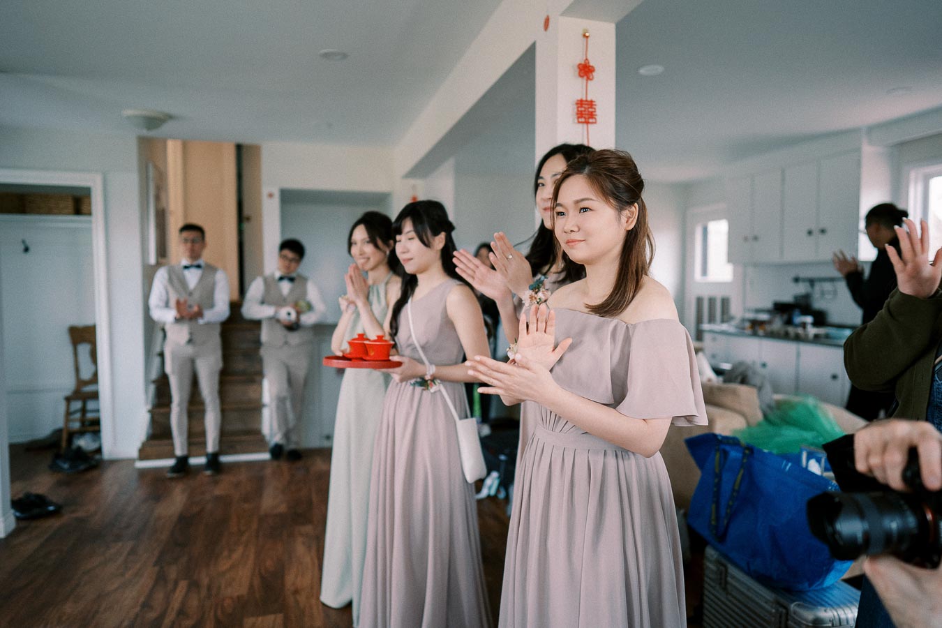 A group of women in pastel-colored bridesmaid dresses clapping during a wedding tea ceremony, surrounded by wedding party members in a bright, modern living room.