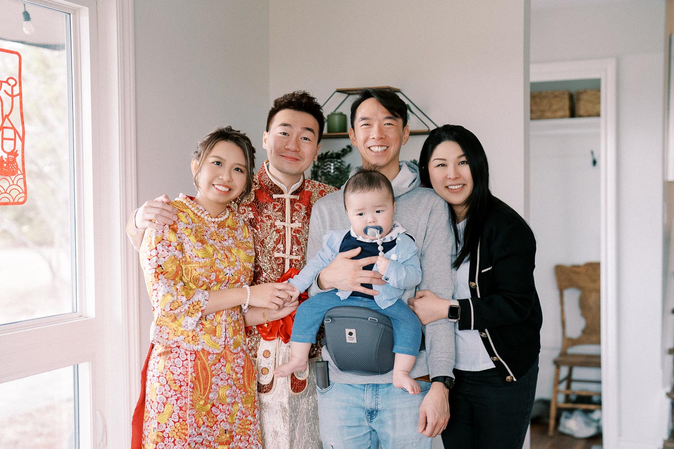A group of smiling people dressed in colorful traditional attire, standing together indoors, with a baby held in a front carrier.