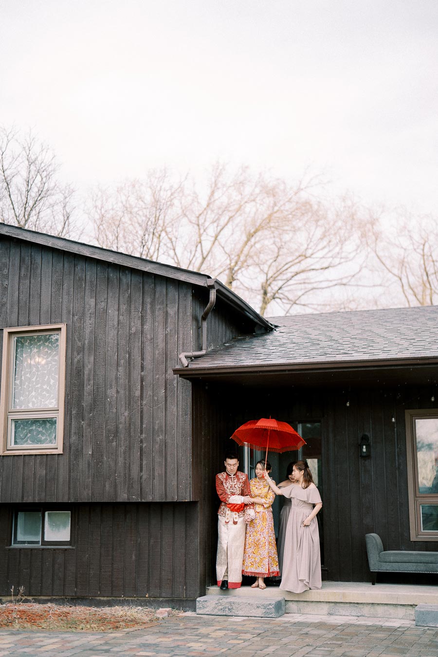 A couple in traditional attire stands under a red umbrella outside a dark wooden house, accompanied by a woman in a light dress. The scene is serene and set against a backdrop of bare trees.