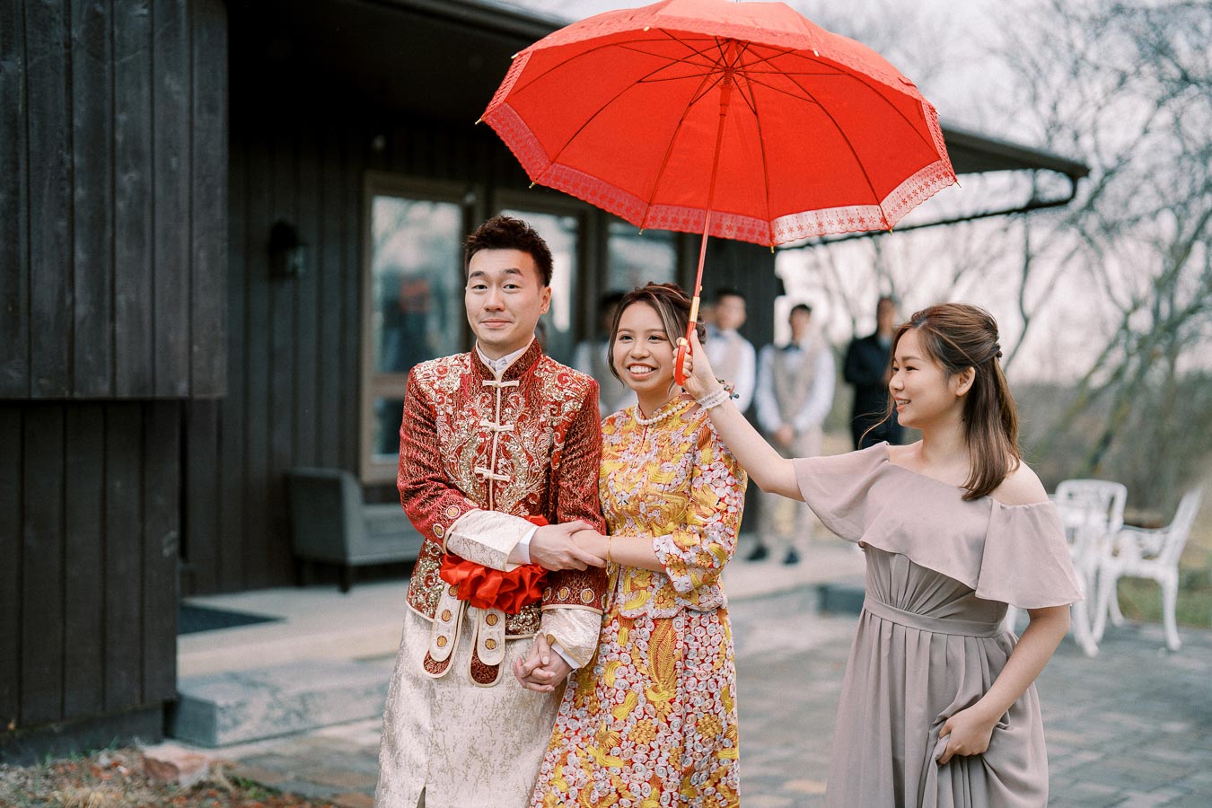 A couple in traditional attire smiling under a red umbrella, with a friend holding it, during an outdoor wedding ceremony.