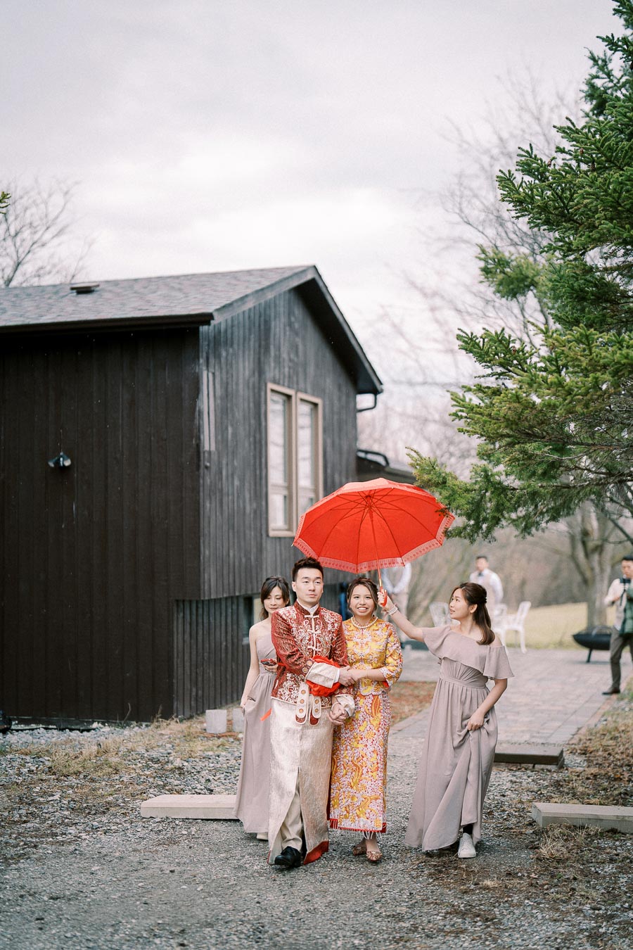 A couple in traditional attire walking outdoors, accompanied by bridesmaids holding a red umbrella, near a rustic wooden building, surrounded by trees and natural scenery.
