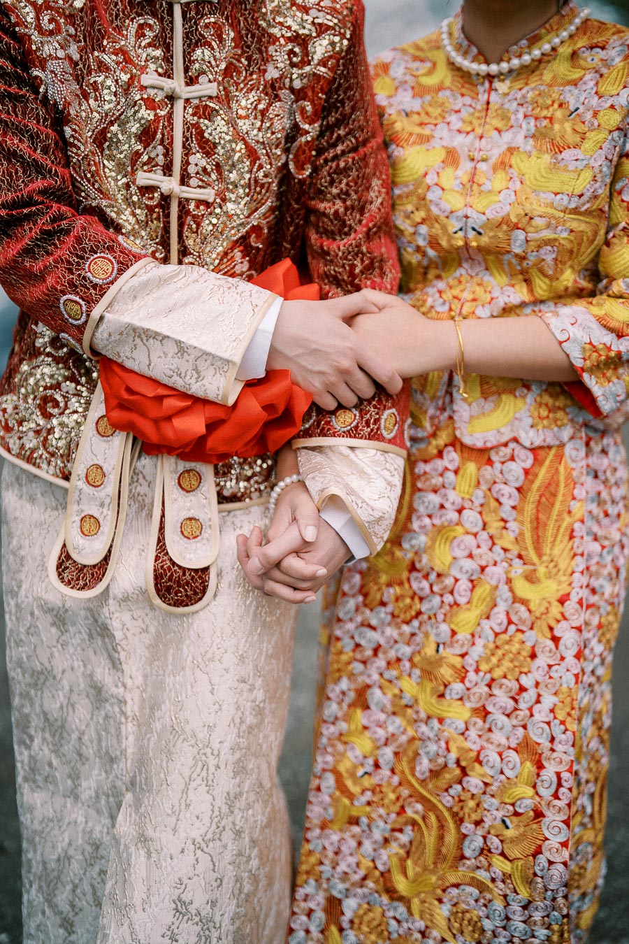 A couple wearing traditional Chinese wedding attire, featuring intricate red and gold embroidery. The bride and groom are holding hands, symbolizing unity and love.