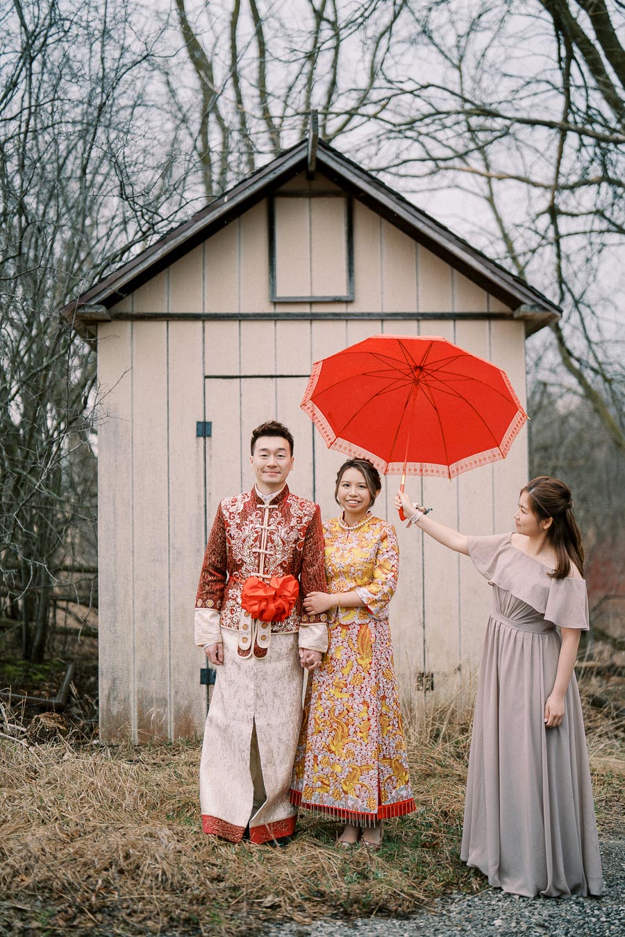 Traditional wedding couple posing in colorful attire with a red umbrella, accompanied by a bridesmaid in a grey dress, standing in front of a rustic wooden shed with bare trees in the background.