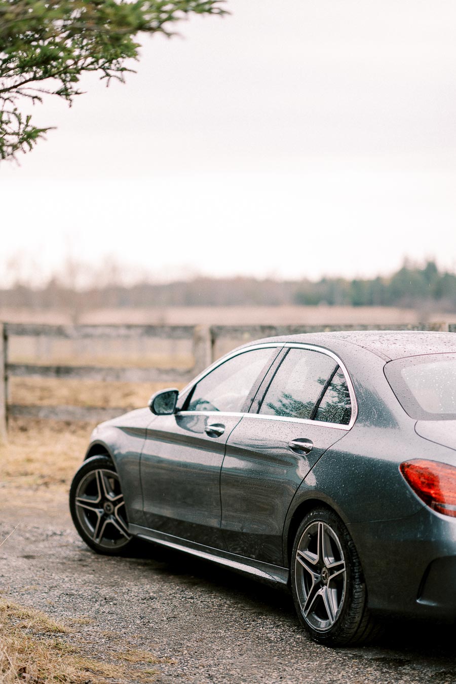 Luxury sedan parked on a rural gravel road with raindrops on the sleek gray exterior and blurred background of wooden fence and trees.