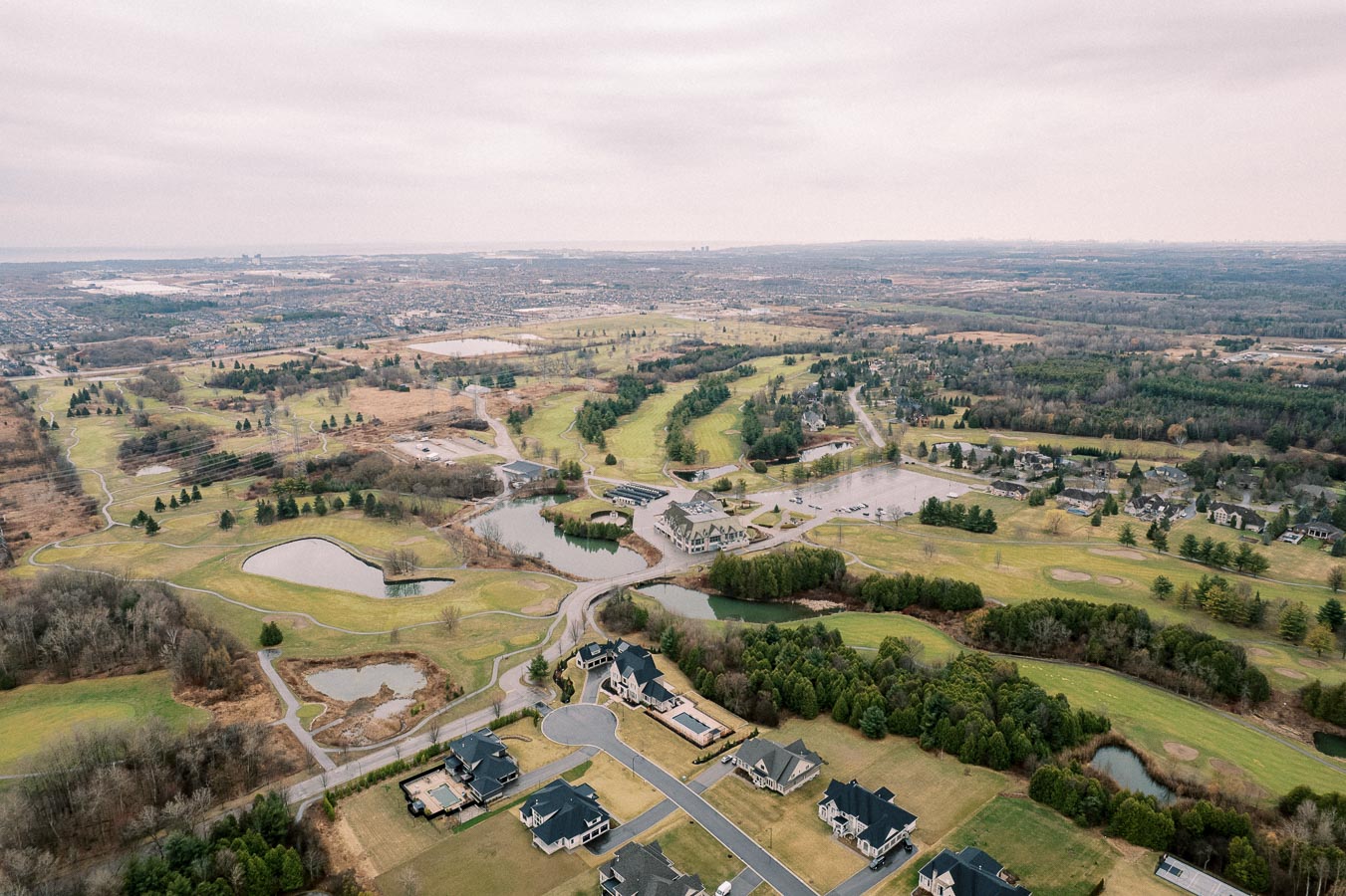 Aerial view of a sprawling suburban landscape with lush green golf courses, residential homes, winding roads, and small lakes on an overcast day.