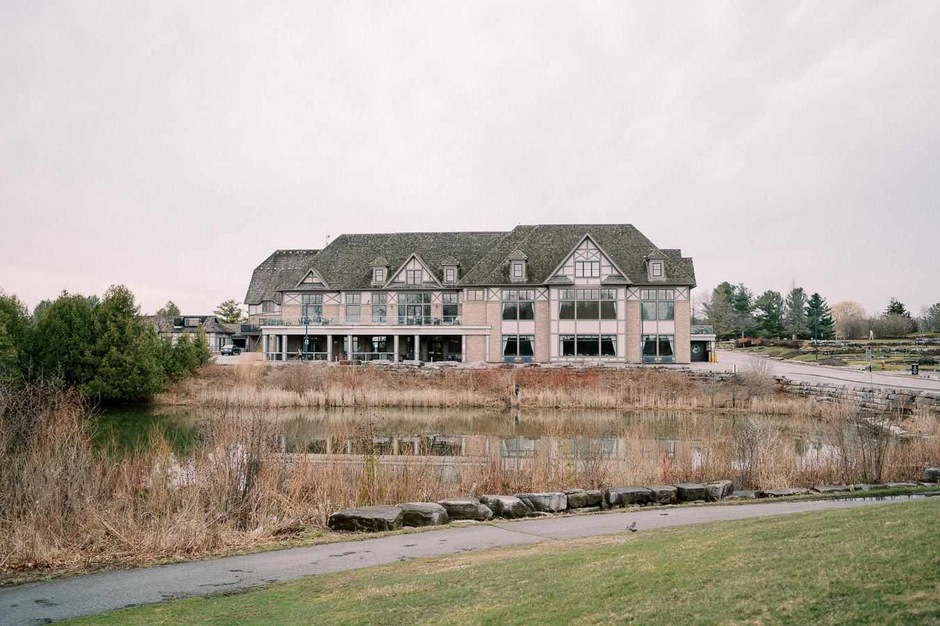Elegant countryside inn with Tudor-style architecture overlooking a tranquil pond, surrounded by lush greenery and a scenic landscape.