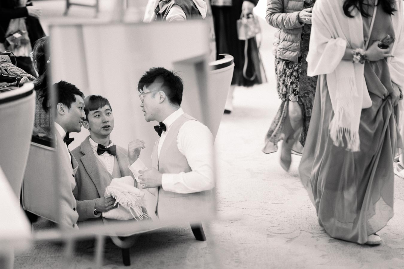 Black and white photo showing a reflection of three formally dressed men engaged in conversation, with people in elegant attire walking past in the foreground, possibly at a wedding or formal event.
