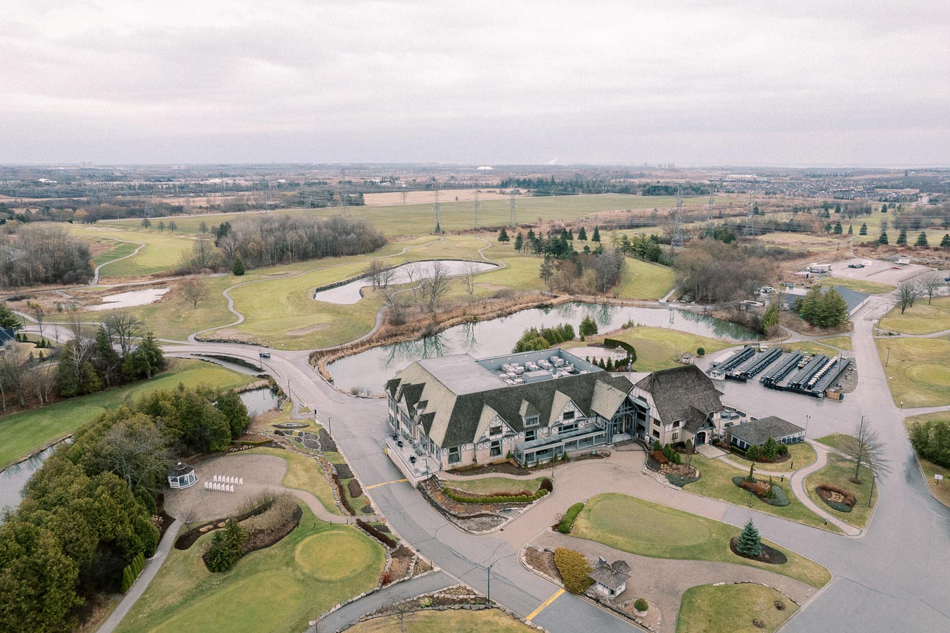 Aerial view of a sprawling golf course featuring a large clubhouse surrounded by lush green fairways, winding paths, and small ponds, set against a backdrop of open countryside under a cloudy sky.