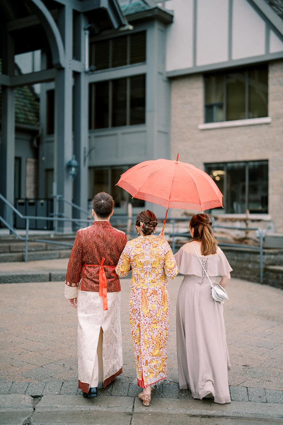 Three people in traditional attire walking towards a building, with one holding a red umbrella on a rainy day.