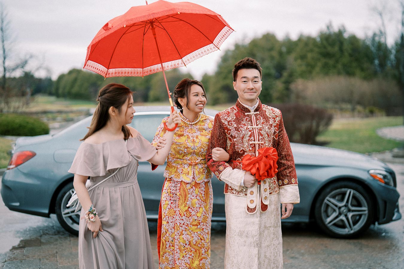 Traditional wedding ceremony with bride in vibrant red and gold attire holding a red umbrella, next to groom in ornate red and white outfit, accompanied by a bridesmaid in a grey dress, standing in front of a luxury car.