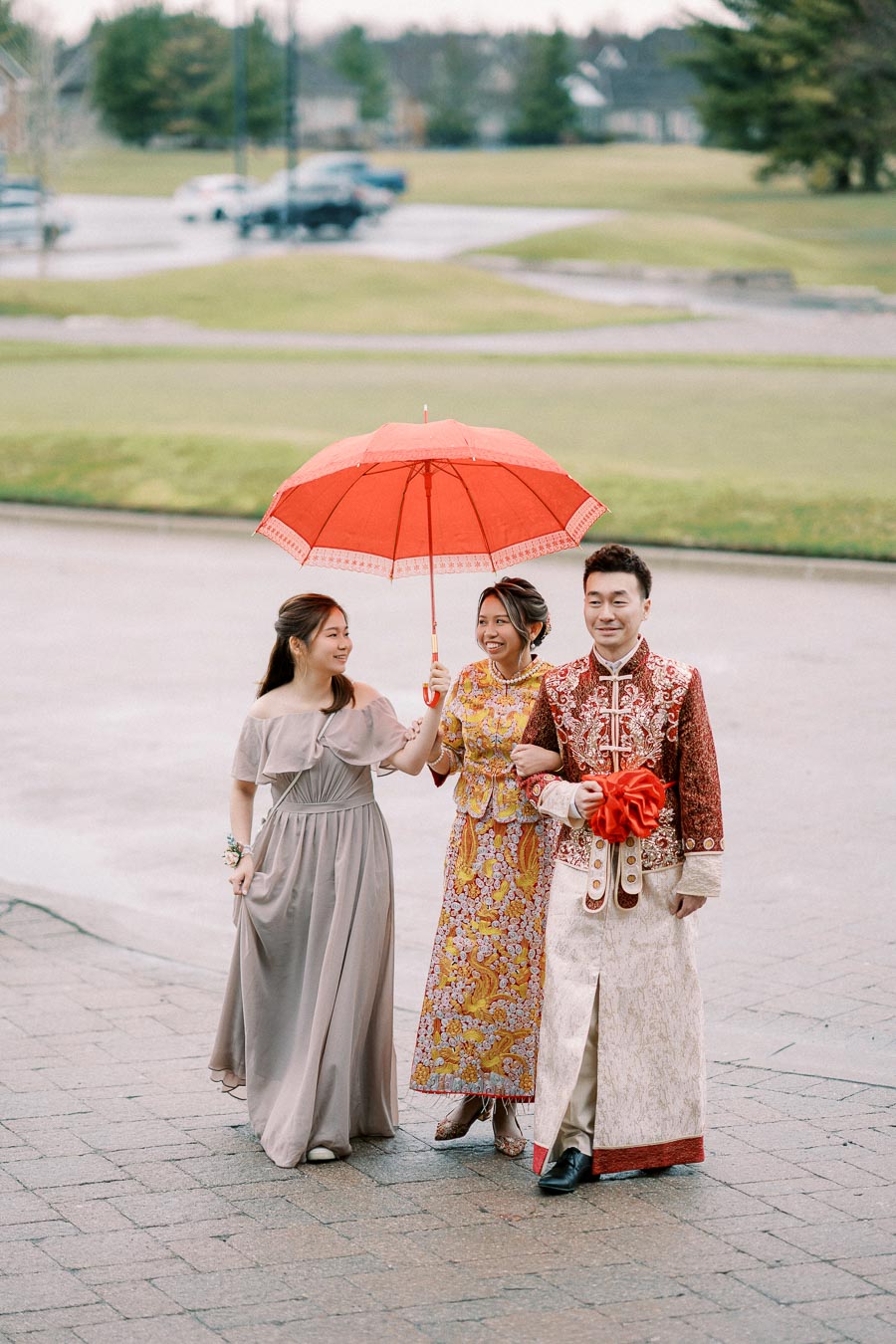 Three people dressed in traditional attire walk together under a vibrant red umbrella on a cloudy day.