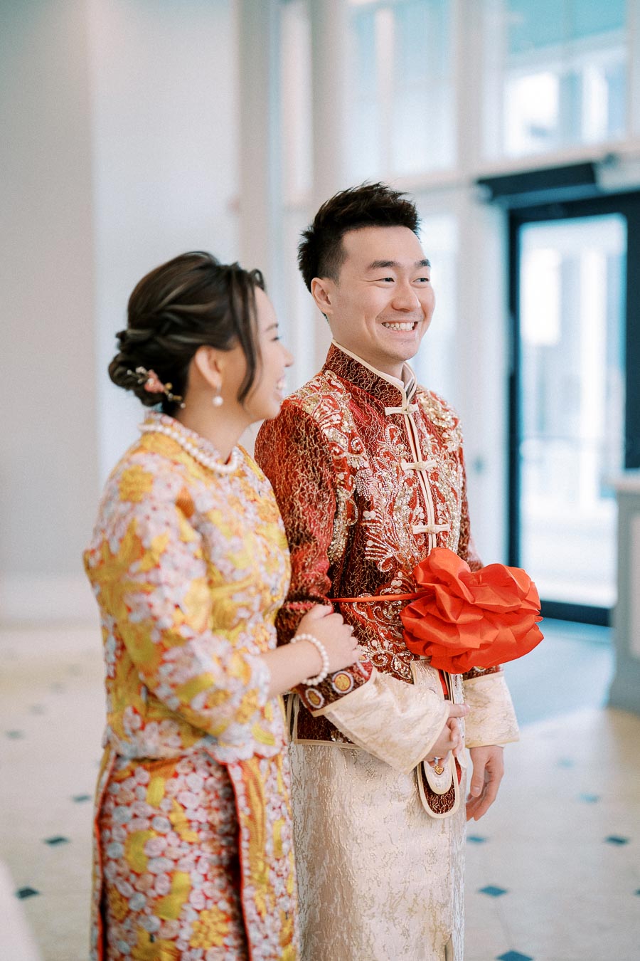 Couple in traditional Asian wedding attire smiling at a ceremony, with intricate red and gold embroidered outfits showcasing cultural elegance indoors.