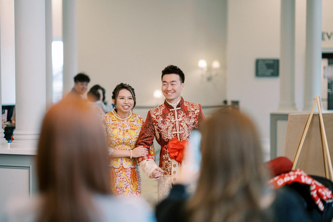 A couple dressed in traditional Chinese wedding attire, smiling and walking arm in arm indoors, with blurred guests in the foreground, capturing the joyful and culturally rich moment.